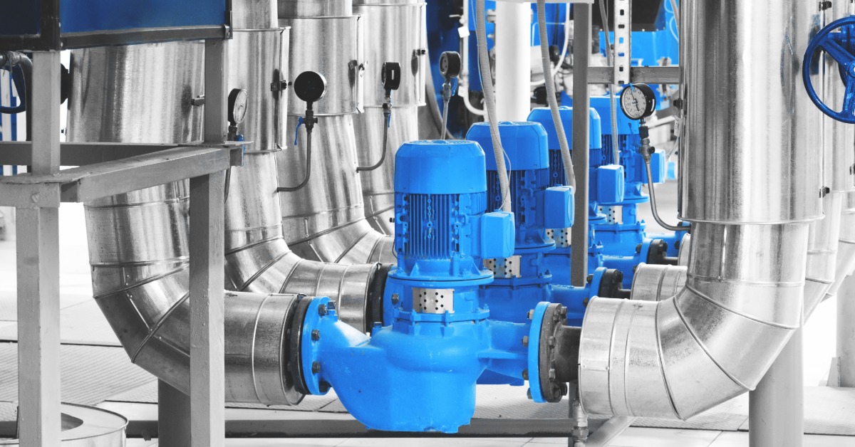 A lineup of several water pumps in a boiler room. There are large silver pipes coming out from each water pump.