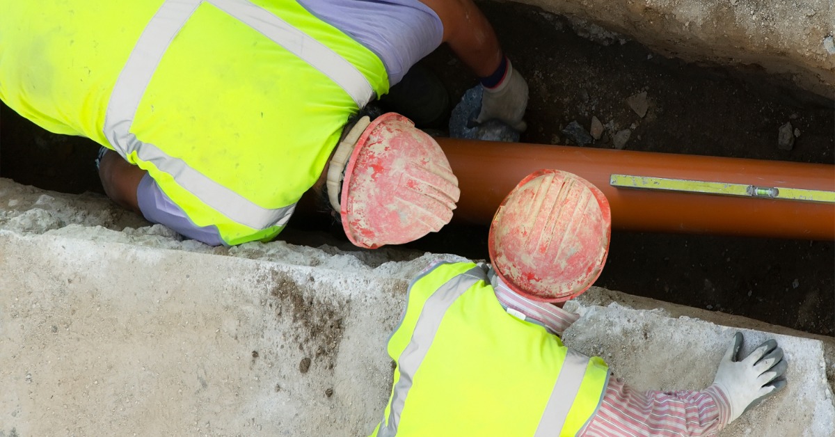 Two construction workers working around an orange sewage pipe that has been uncovered from the ground.
