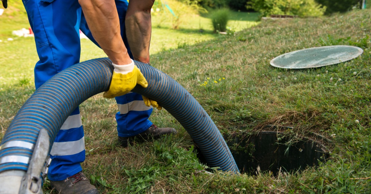 A sewage professional in blue coveralls putting a large hose down into a hole in the ground where a septic tank is.