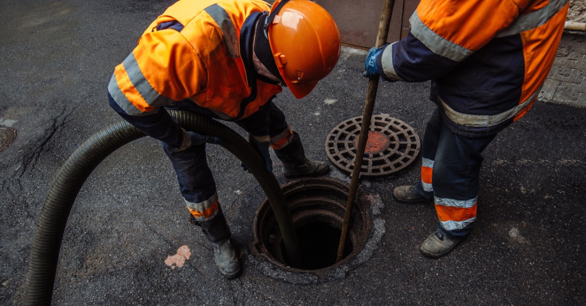 Two sewer workers in orange safety vests working together to unclog and clean a sewer opening in the ground.