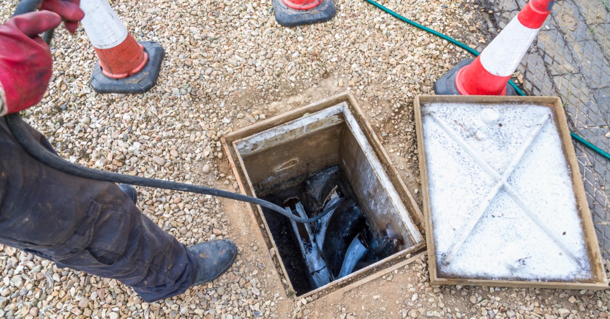 A man in black boots using a small hose to clean out a sewer pipe that is clogged and below a rectangular manhole.