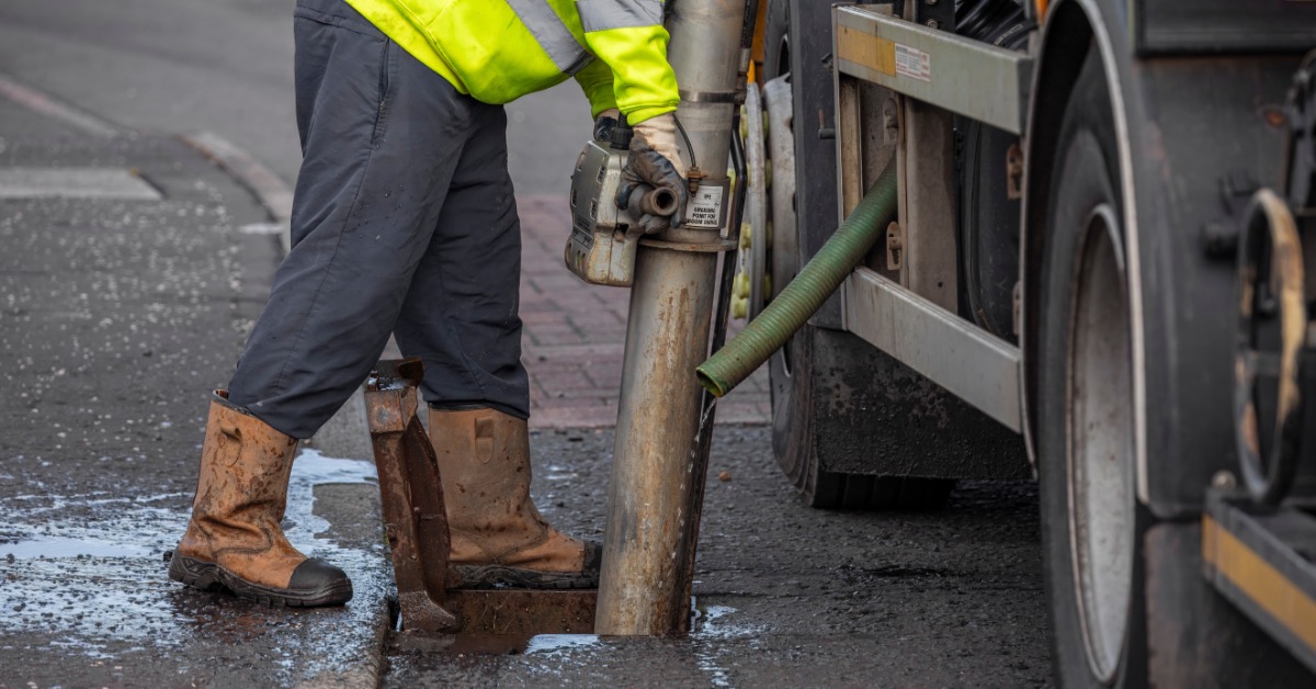 A worker in a yellow hi-vis vest using a large metallic pipe to clean out a sewer drain in the ground.