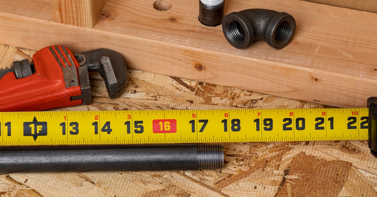 A black steel pipe fitting rests next to a black pipe on a board next to a tape measure, wrench, and longer pipe.