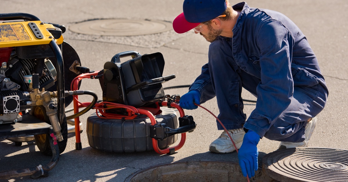 A man kneels down in front of a monitor as he feeds a camera inspection line into the opening of a wide drain system.