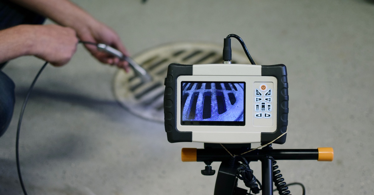 Focus on a small monitor hooked up to a sewer camera inspection line as a person feeds it into a grated drain pipe.