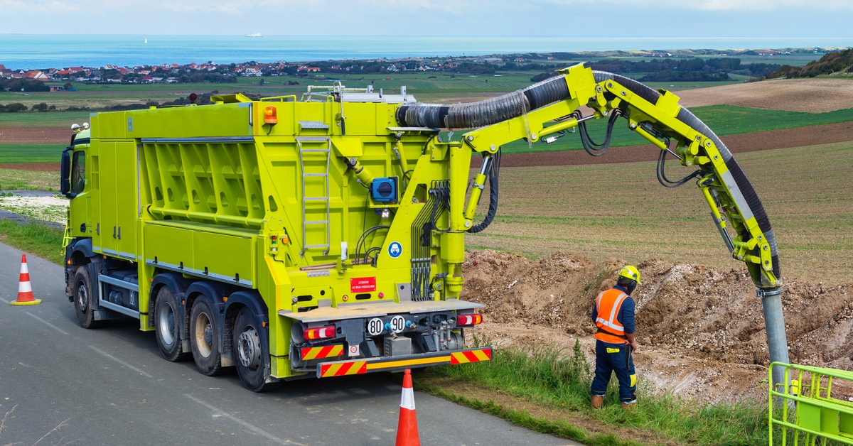 A hydrovac truck parked off to the side of a road. A man in a vest and hard hat examines the area the boom is directed to.