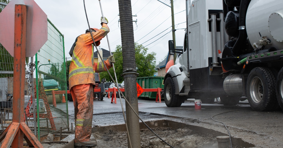 A man wearing an electric orange jumpsuit operates hydrovac equipment at a carved-out portion of pavement near a road.