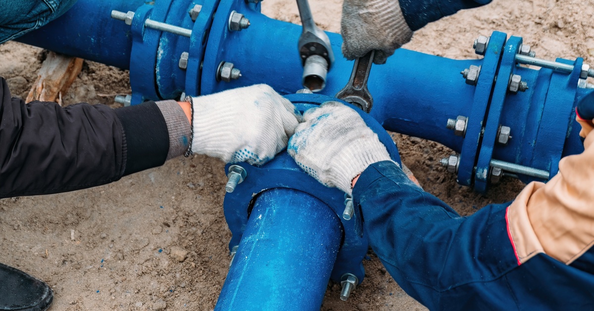 Two people wearing gloves are holding and repairing large, blue pipes with a wrench and a torque wrench.
