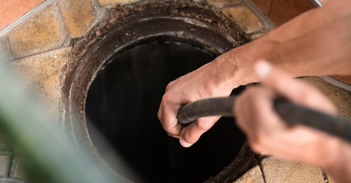 A close-up of a person feeding a black hose into an open drain pipe in a portion of a brown tiled floor.