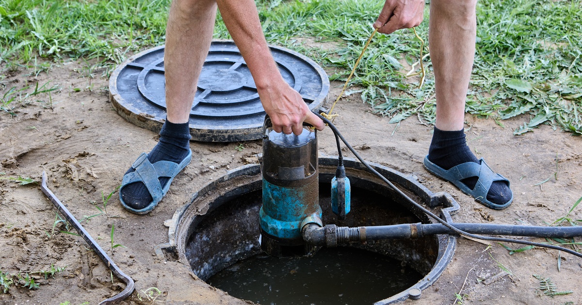 A person wearing sandals and black socks holds a rusted blue water pump over an open hole in the ground.