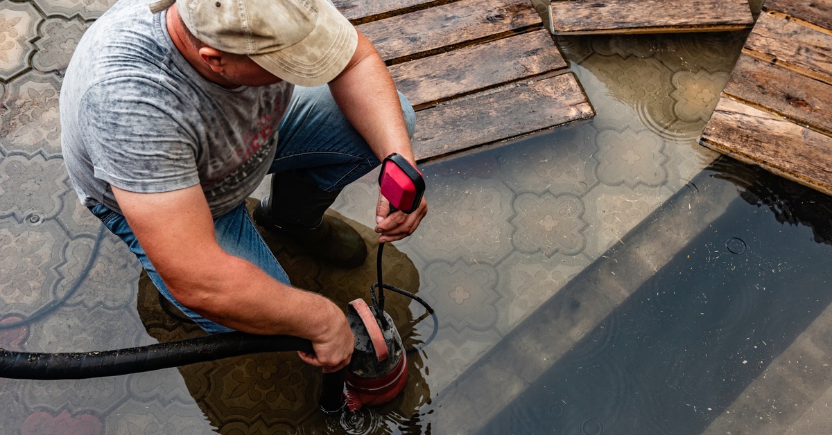 A man in a dirty T-shirt uses a submersible pump in a pool of water with wooden boards floating nearby.