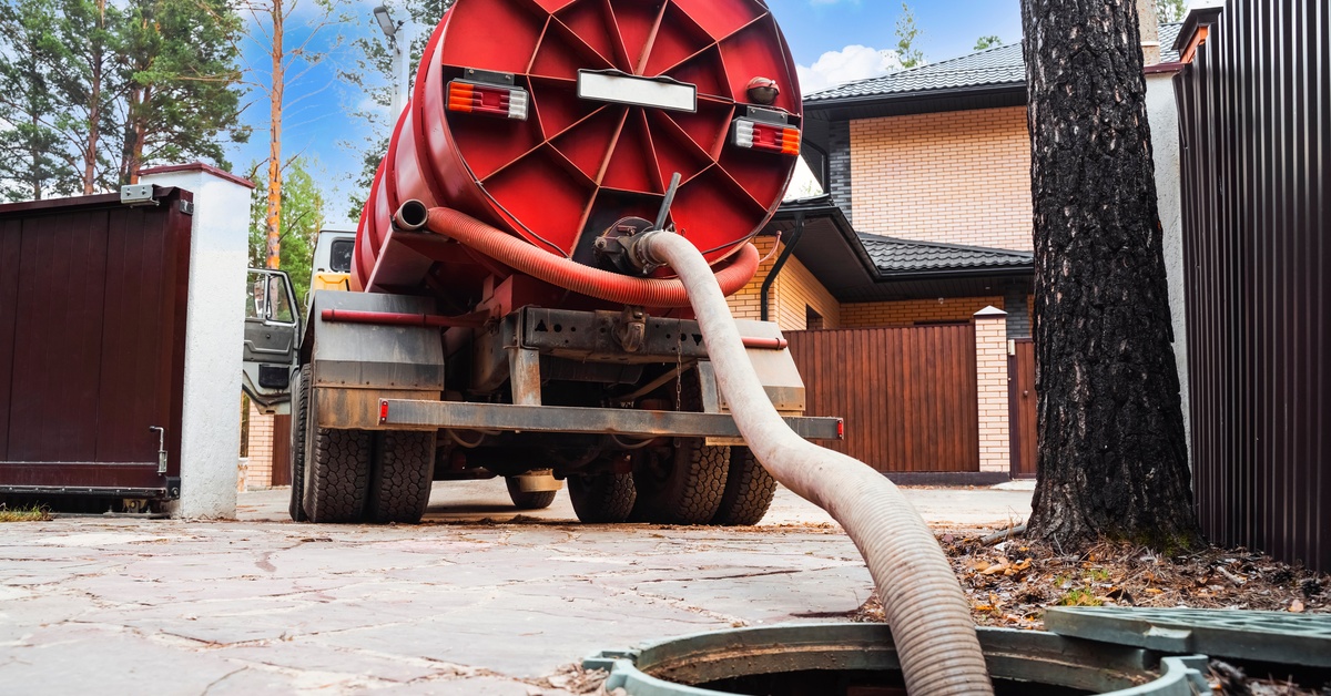 A truck with a red tank and an attached hose that extends into an open sewer entrance in a suburban area.