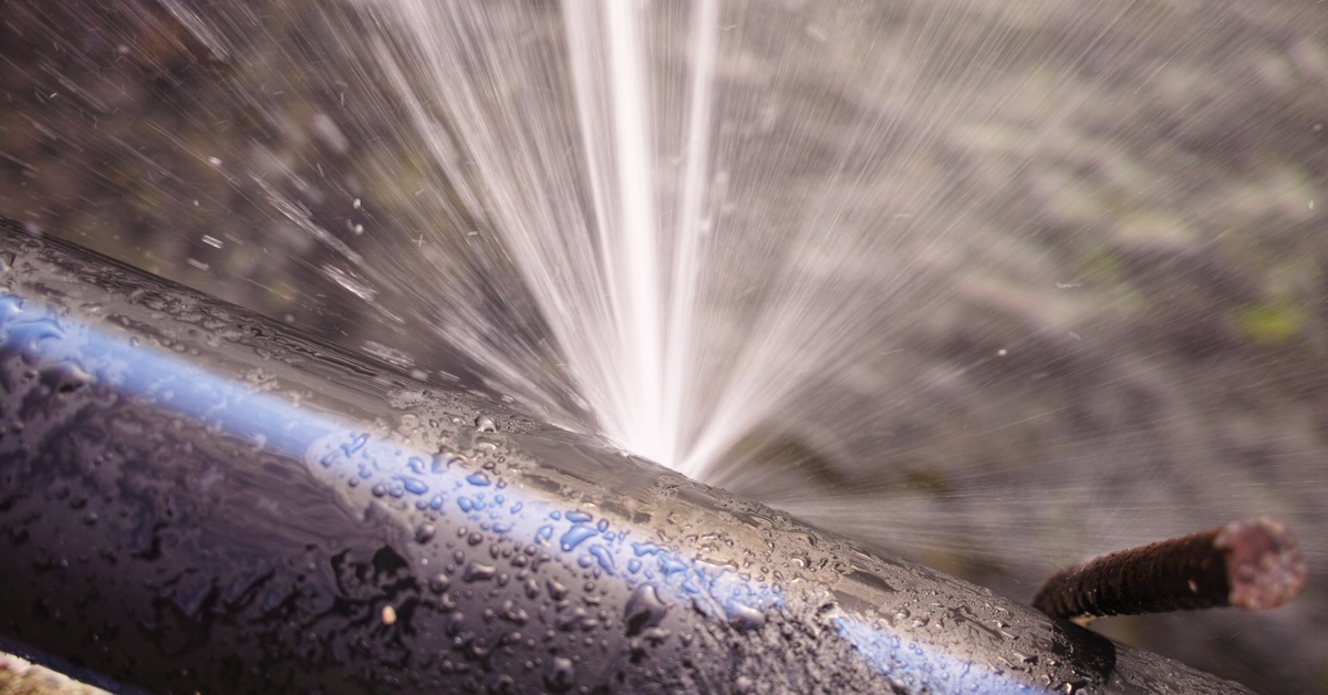 An old pipe with a leak that powerfully sprays water in multiple directions, coating its metal surface.