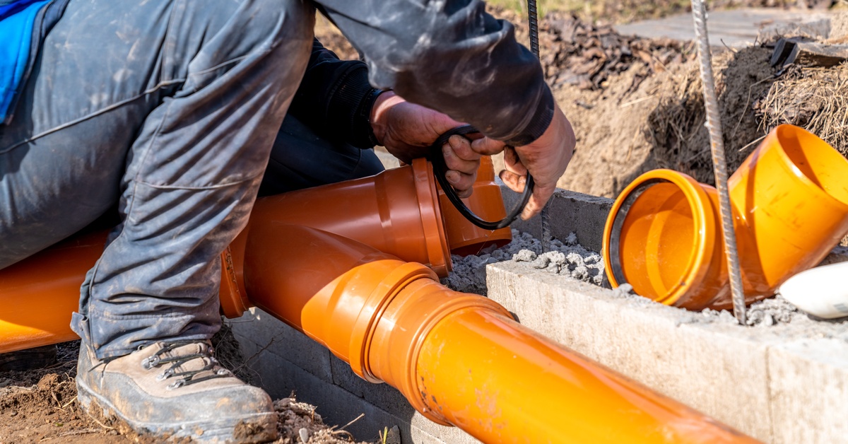 A person in work boots, jeans, and a blue shirt crouches over a disassembled orange pipe in a ditch.