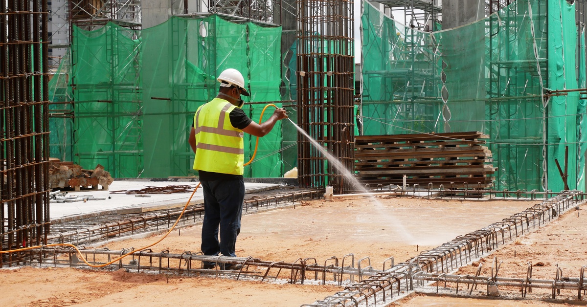 A person wearing a hard hat and neon vest spraying a high-pressure hose at the ground on a construction site.