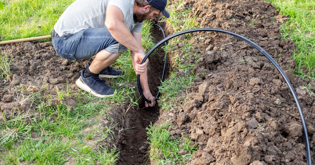 A man shifts a long hose inside an irrigation trench in between mounds of dirt and rock in a grassy field.