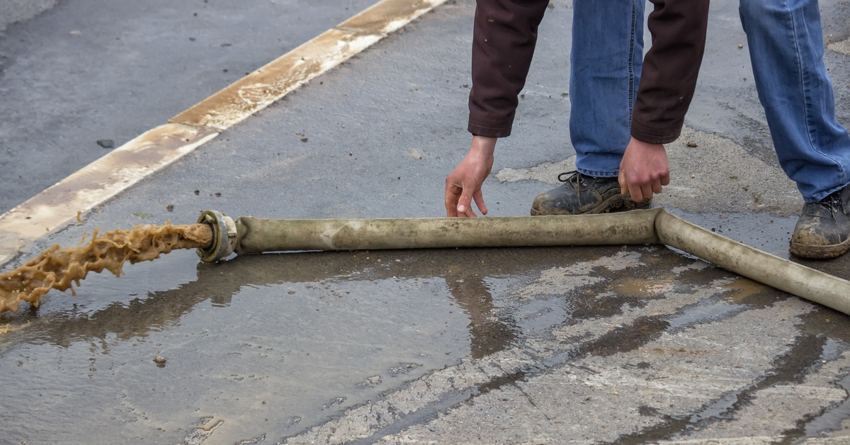 A person operating a hose that ejects brown water out of its opening while on wet asphalt on a street.