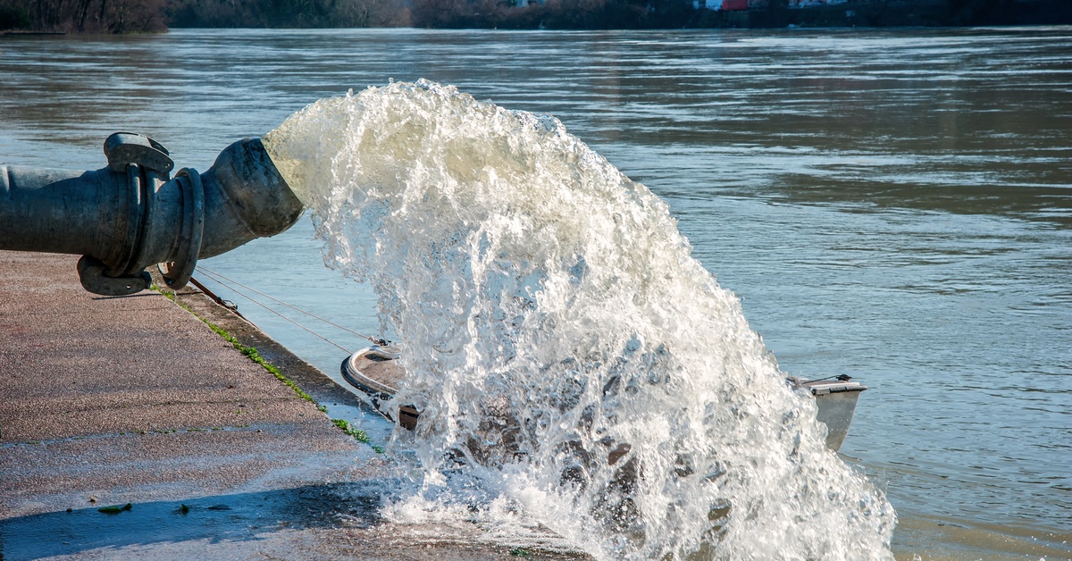 A large hose ejecting water into a small lake on a wet concrete dock with trees and houses in the distance.