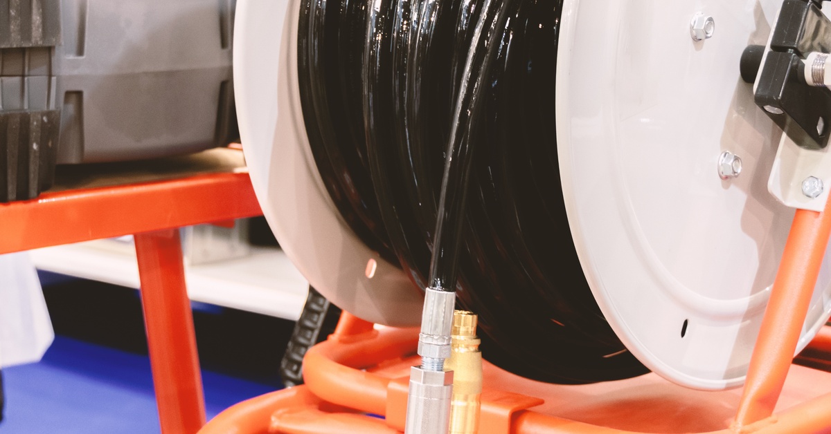 A black industrial hose connected to a nozzle and coiled onto a large white reel. The reel rests on a structure with orange bars.