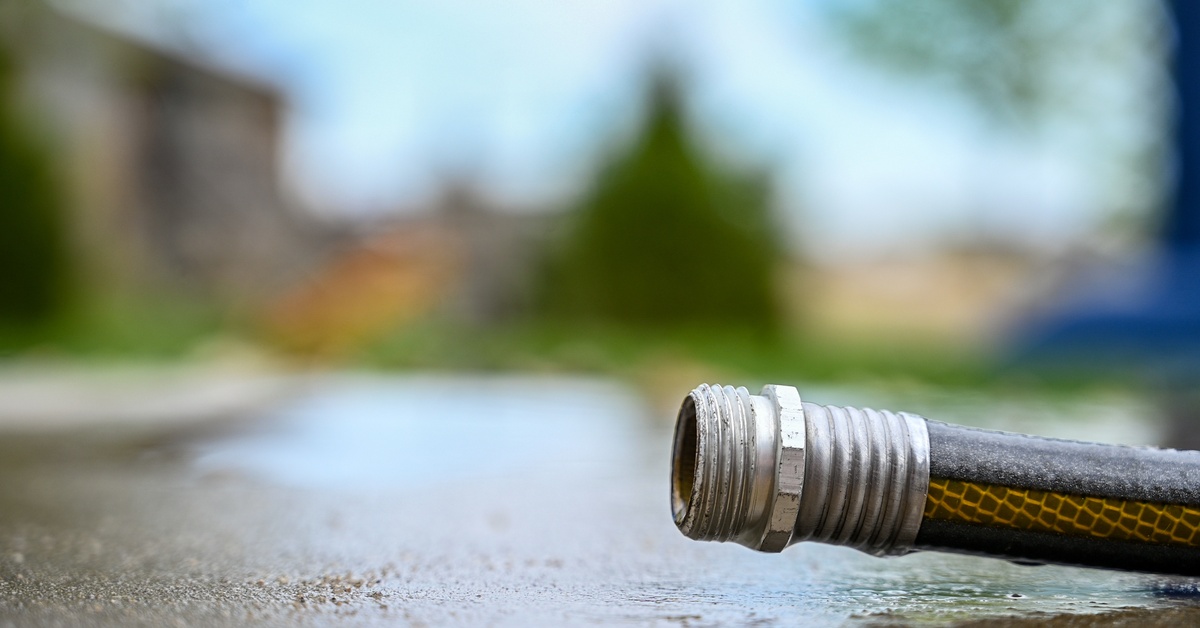 The metal tip of a hose made of black and yellow material lying on wet asphalt with a yard in the background.