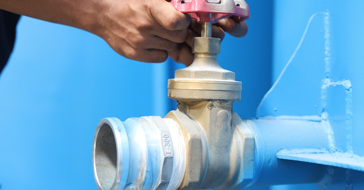 A person turning the red valve on top of a brass ball valve connected to the side of a blue metal surface.