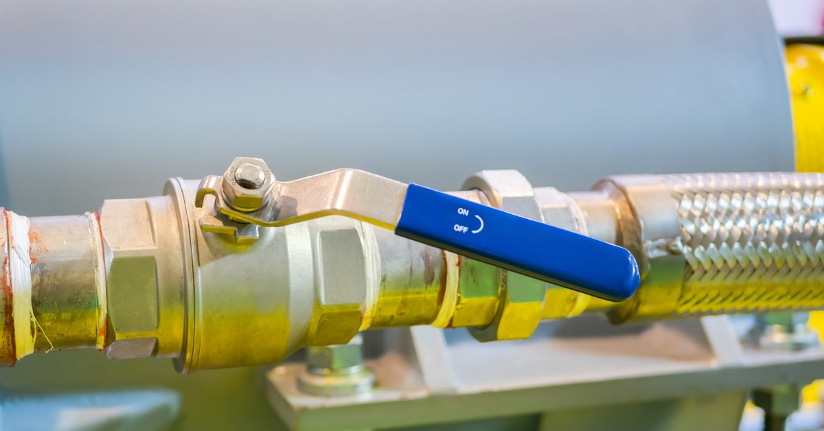 A brass ball valve with a blue on-and-off handle connected to a braided pipe next to a large tank in a facility.