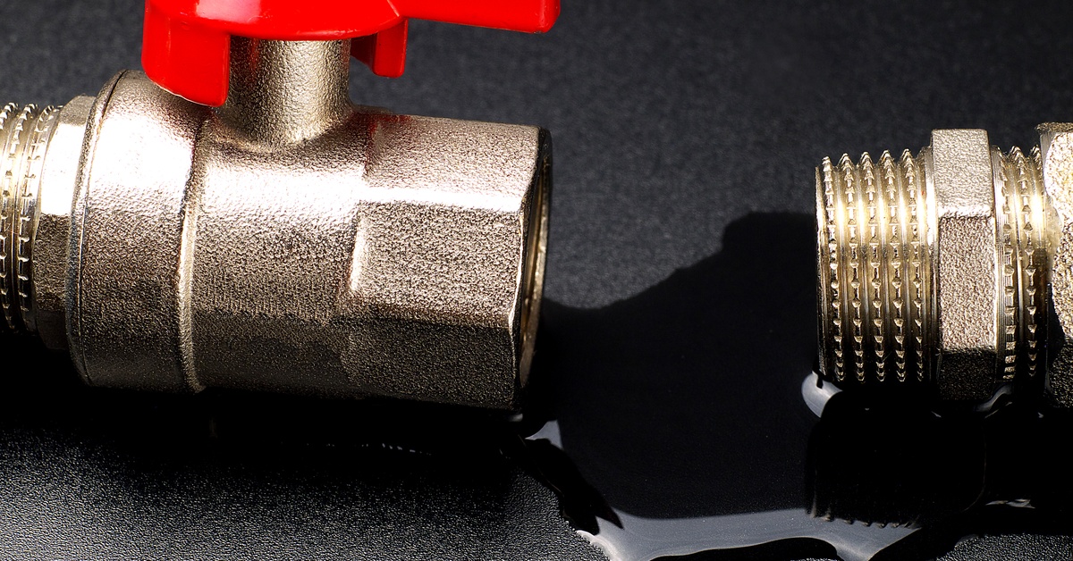 Water drips out and forms a puddle between a disconnected ball valve and fitting laying on a plain black surface.