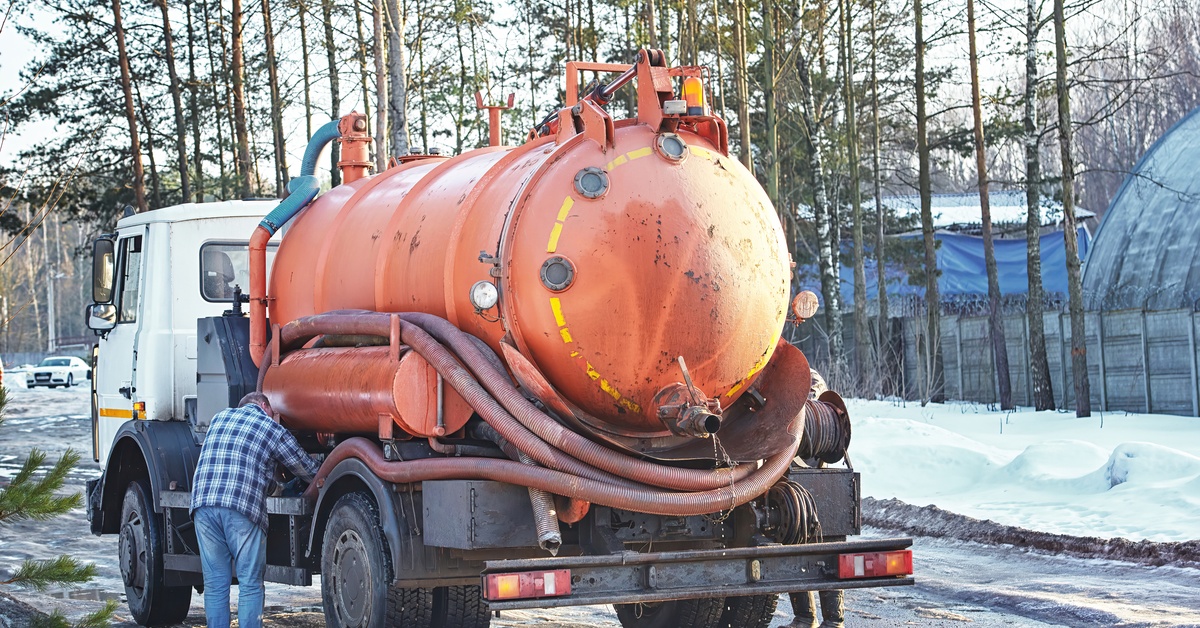 An orange vacuum truck with a leaking spout is parked on the side of a snowy road while two people inspect the hoses.