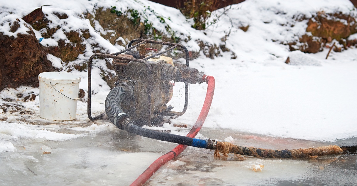 A water pump connected to two hoses sits on the ground outside near melting snow. A white bucket is nearby.