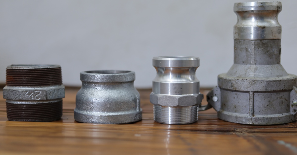Four Camlock fittings including gaskets, adapters, and pipe reducers lined up next to each other on a wooden surface.