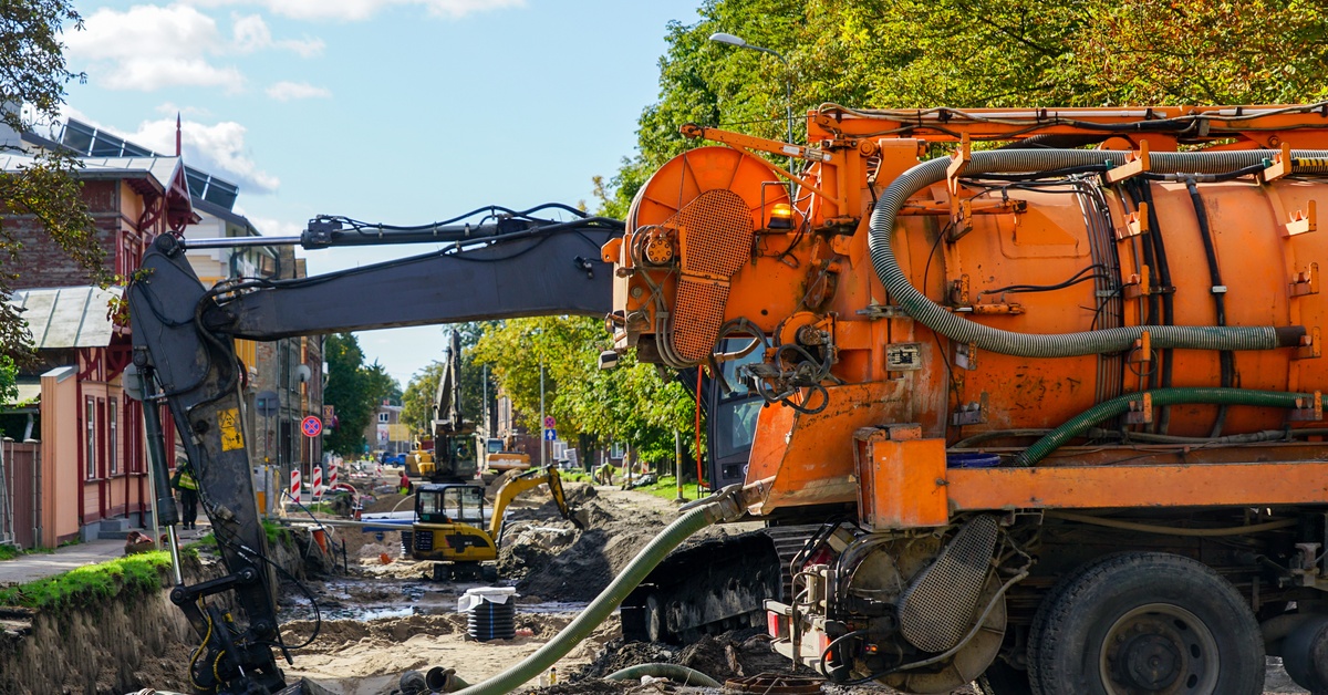 An orange vacuum truck and a black backhoe performs construction work on a street that extends into the distance.