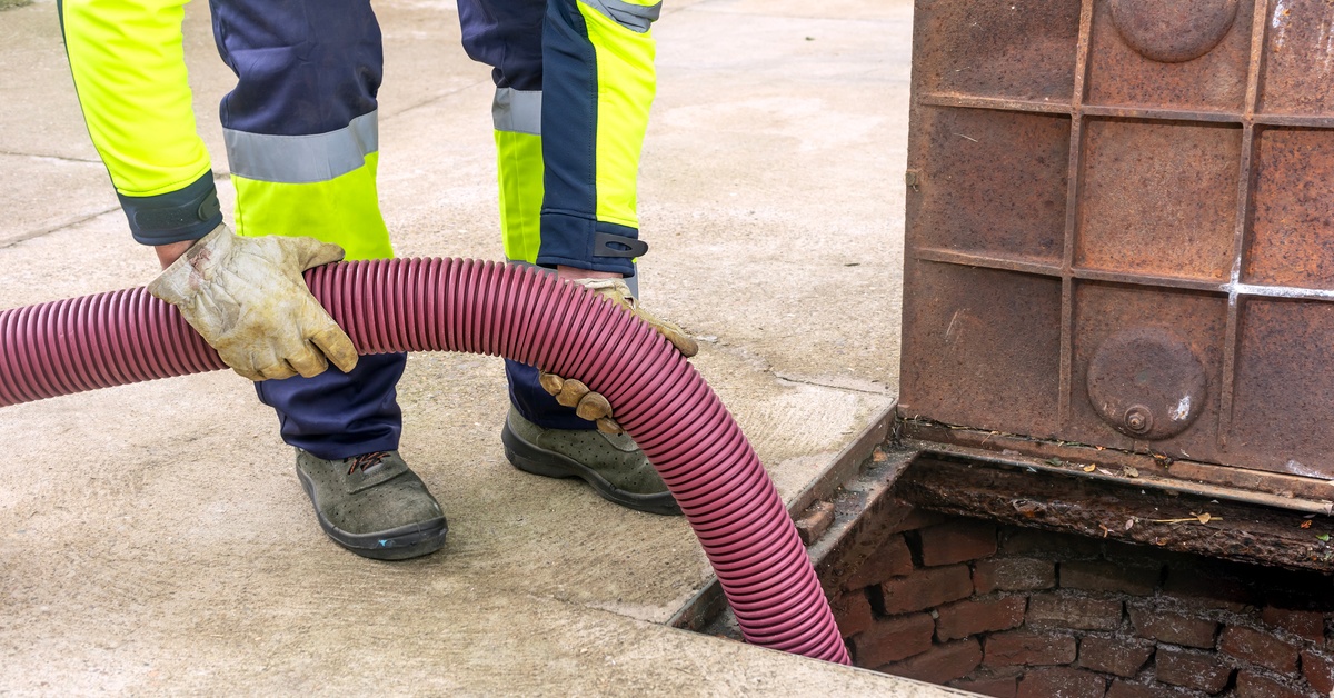 A person wearing dirty white loves and high-visibility protective clothing places a thick, red hose into a drain pipe.