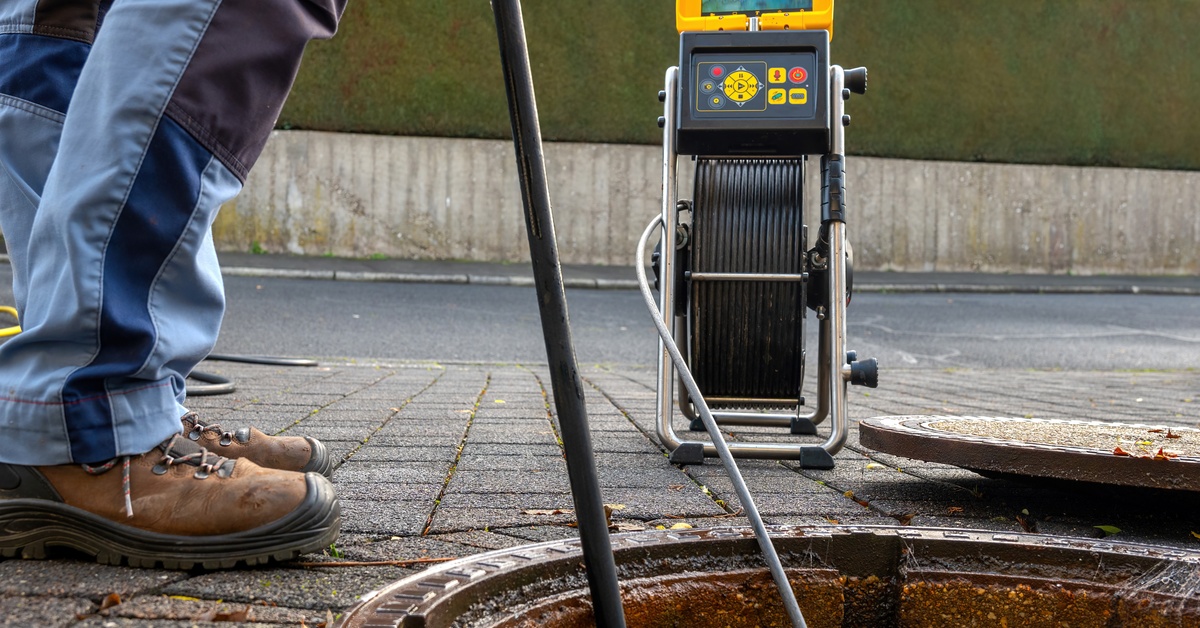 A person standing by an open maintenance hole with the cable of a sewer camera being fed into it as the nearby device provides a feed.