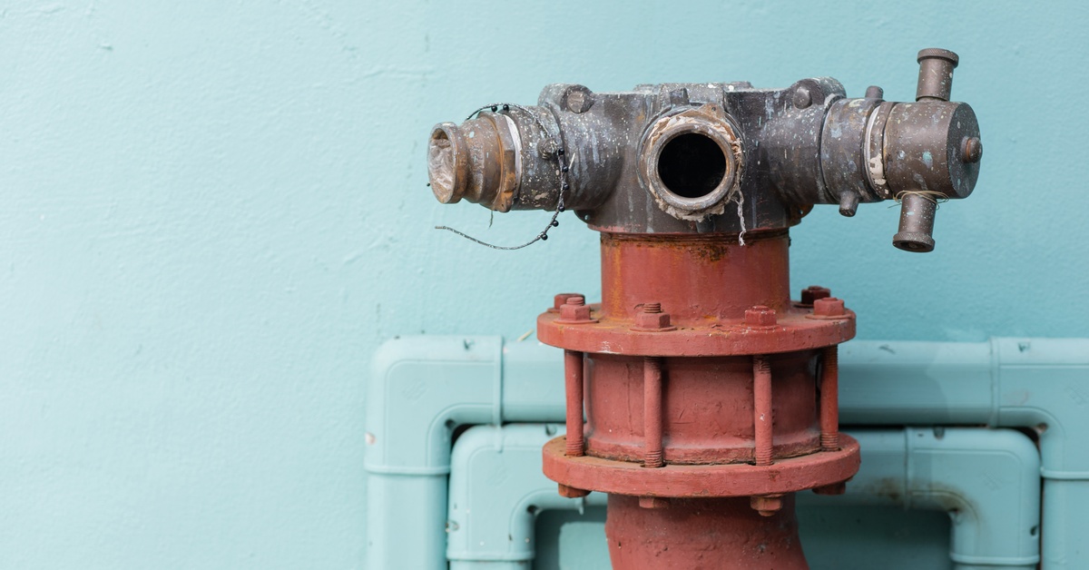 A red-and-brown hydrant with three heads containing various openings and adapters placed against blue wall with piping.